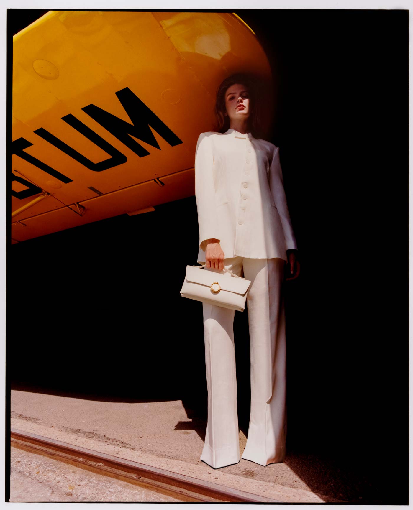 Person in a white suit holding a mlouye handbag with a yellow vehicle in the background
